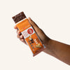 A dark chocolate peanut butter bar sticking out of the orange packaging being held by a woman's hand.