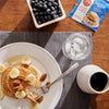 Plate of pancakes with sliced banana and pecan toppings, a glass of ice water, syrup, blueberries, and a single serving package of the Oats and Walnuts Pancakes on a wood table top