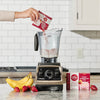 Hand pouring a package of Strawberry Banana Smoothie Mix into a blender with bananas, strawberries, and a 12 pack product box sitting on the counter beside the blender